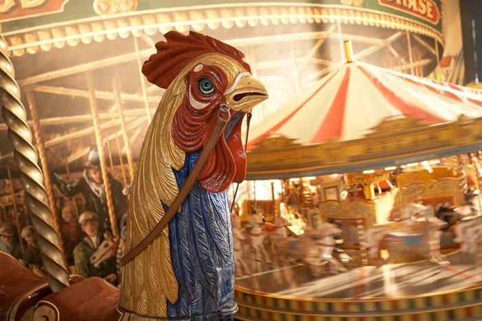 A close-up image of a cockerel galloper from a fairground carousel, in front of a spinning carousel model.