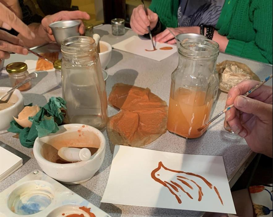 Image of natural pigments in glass jars and closeup of a hand painting a bird.