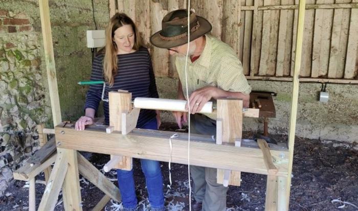 Female student on left. Male tutor wearing hat on right: demonstrating turning wood on a spindle.